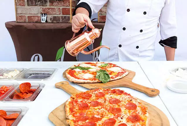 Chef drizzling olive oil over a fresh Margherita pizza beside a pepperoni pizza on wooden peels — Basil Pizza Bar, the site you're on